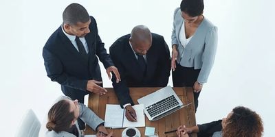 Five business professionals discuss work around a table with documents and a laptop.