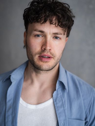 Young man with curly hair wearing a blue shirt and white tank top.