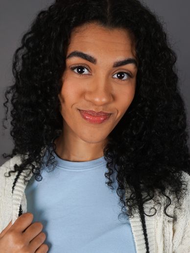 Young woman with curly hair smiling gently, wearing a light blue top and white cardigan.