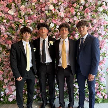 Four young men in suits posing in front of a floral backdrop.