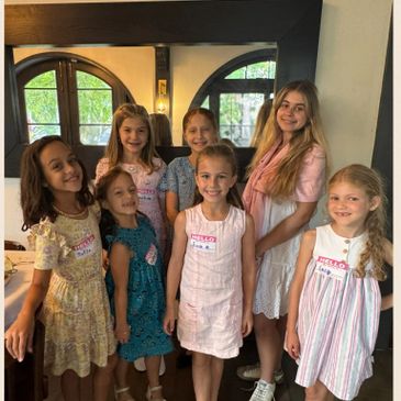 Group of seven young girls smiling indoors, wearing name tags and summer dresses.