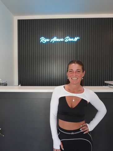 Smiling woman in activewear stands indoors with neon sign.