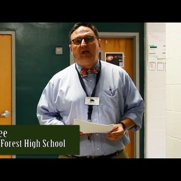 A man with glasses and a bow tie standing in a school hallway, holding papers.