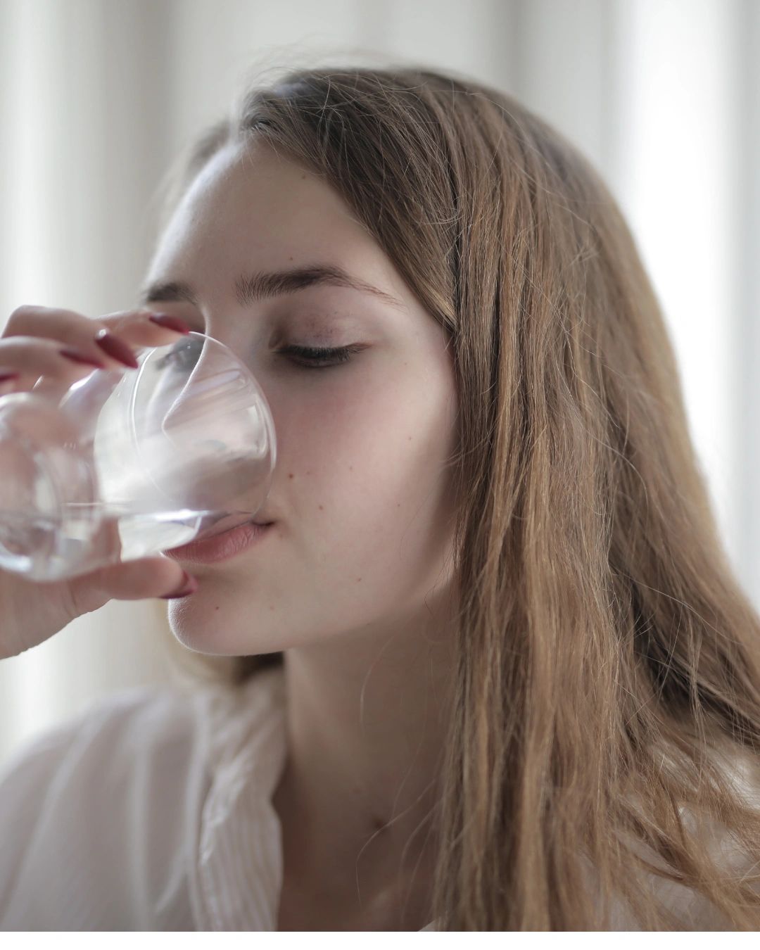 A young woman drinks water from a clear glass indoors.