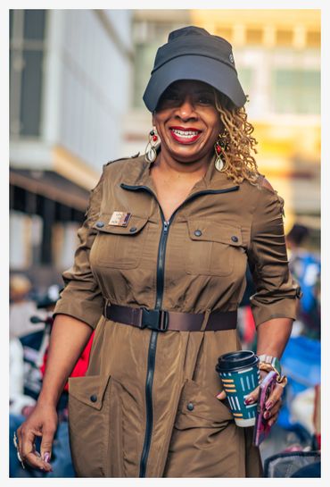 Smiling woman in a brown dress and black cap holding a coffee cup outdoors.