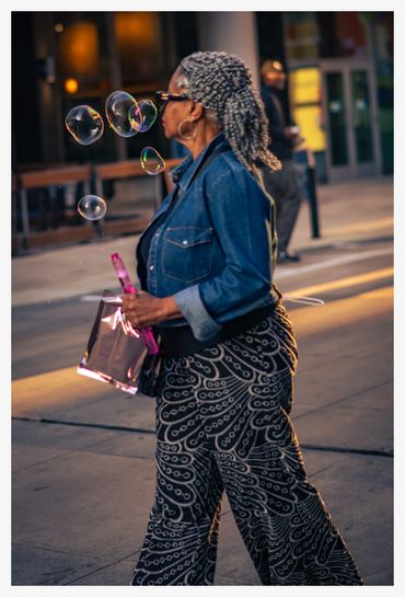 Woman blowing bubbles while walking outdoors in patterned pants and denim jacket.