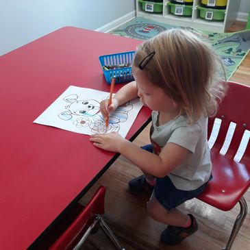 A young child colors a drawing of a bunny and flowers at a red table.