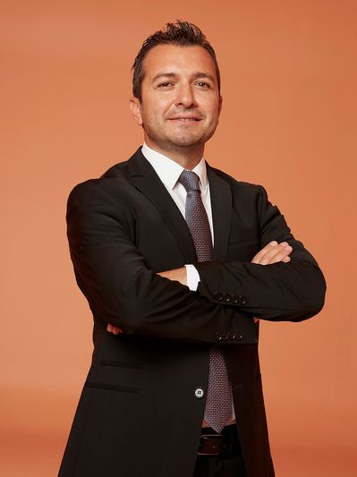 Confident man in a black suit and tie standing with arms crossed against an orange background.