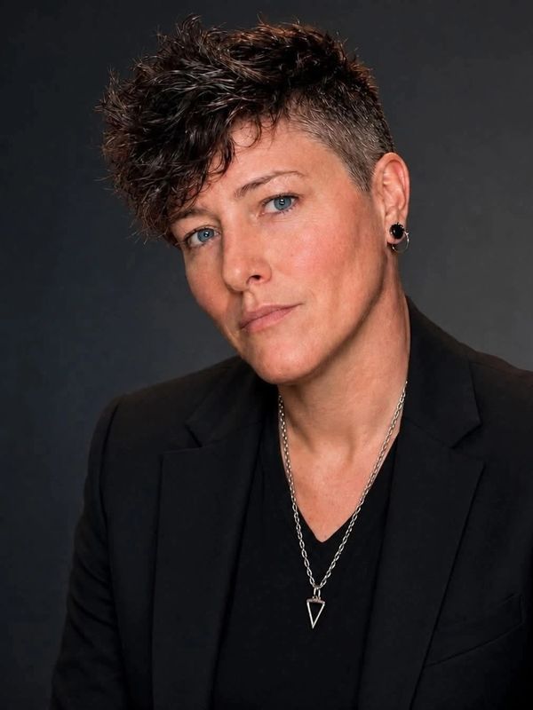 Person with curly short hair and blue eyes wearing a black blazer.