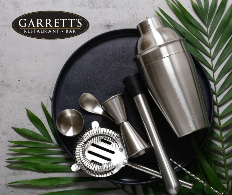 Cocktail-making tools on a black plate with greenery and Garrett's logo.