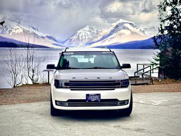Good Vibe Rides White Ford Flex SUV parked by a lake with snow-covered mountains in the background.