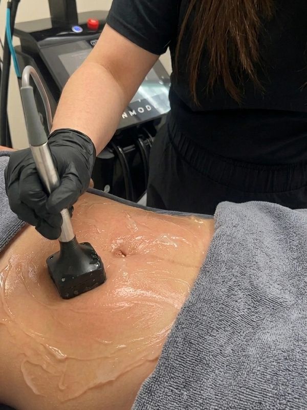 A technician performs a body treatment using a handheld device on a gel-covered abdomen.