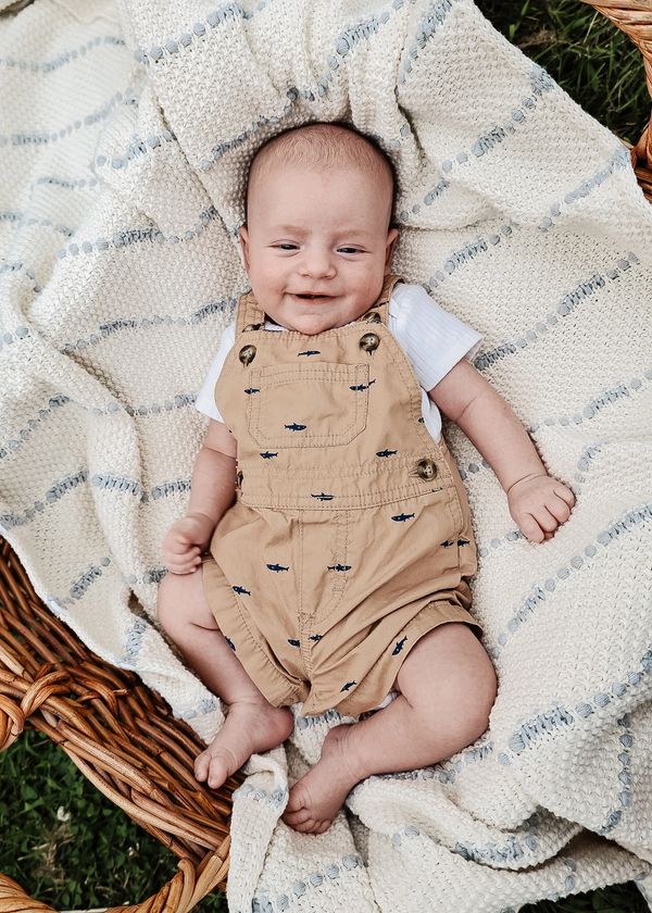 Smiling baby in beige overalls lying on a cozy blanket in a wicker basket.
