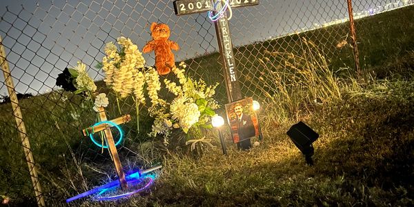 A nighttime memorial with crosses, flowers, a teddy bear, and a photo illuminated by lights.