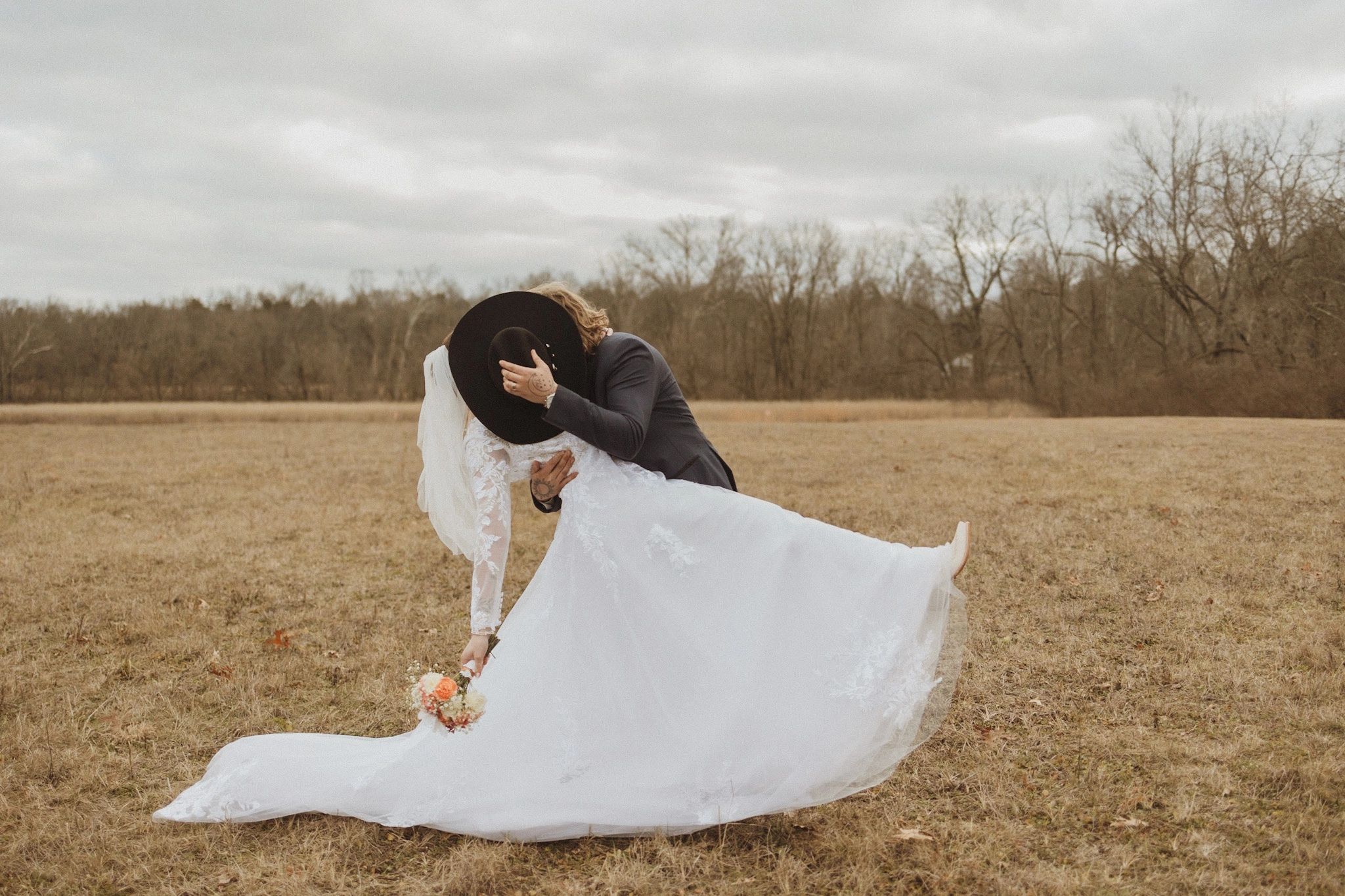 Bride and groom share a kiss in a field, groom hides behind a hat.