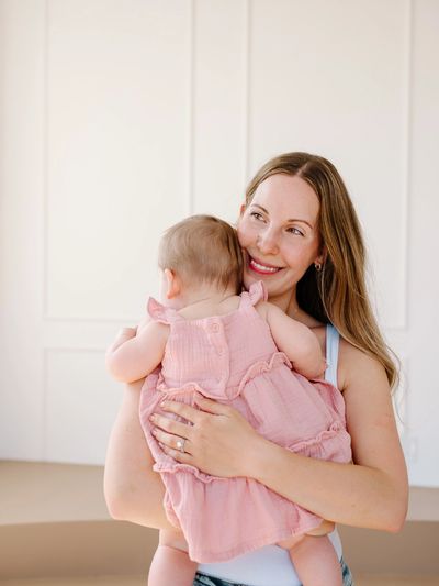 A smiling woman holds a baby in a pink dress indoors.