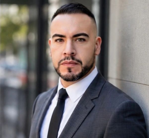 Confident man in a suit posing outdoors against a wall.