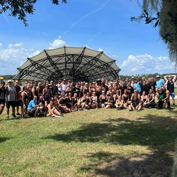 Large group posing outdoors in front of a modern pavilion on a sunny day.