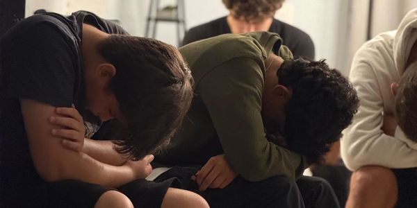 A group of young people sitting with heads down, appearing distressed or reflective.