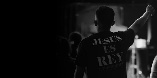 Person raising fist wearing a shirt that says 'Jesús es Rey' in a dark setting.