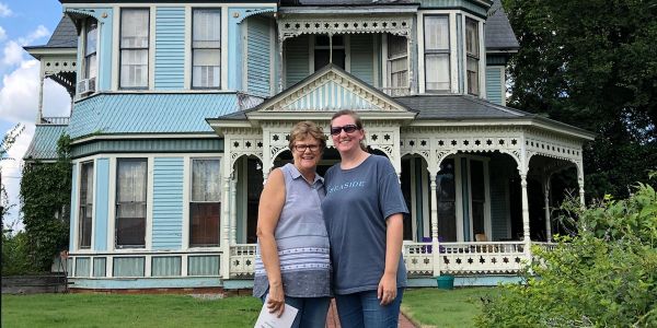 Two smiling women stand in front of a large blue Victorian house.