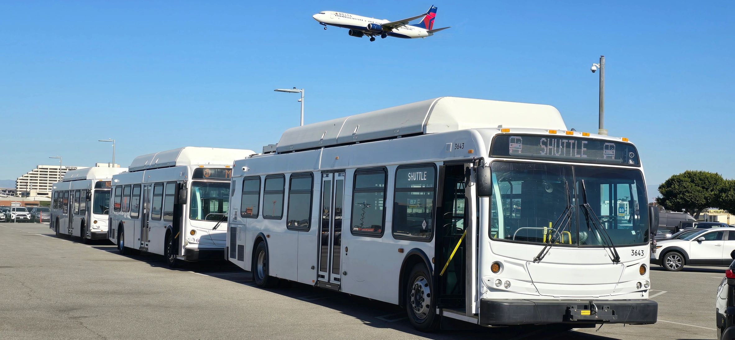 Three white shuttle buses parked in a row with a Delta airplane flying overhead.