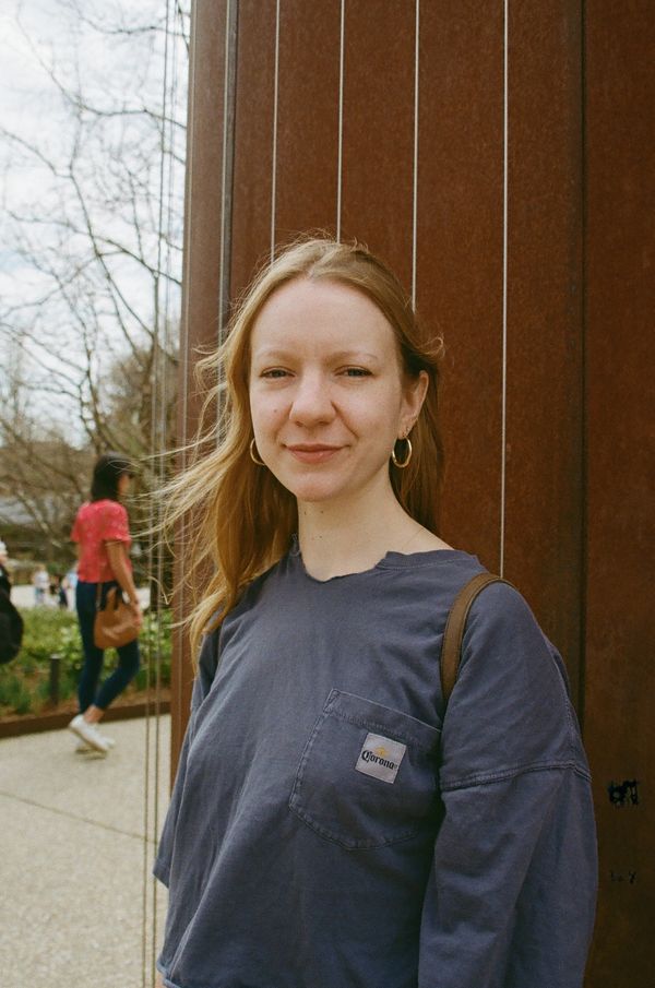 Young woman with long hair stands outdoors near a rust-colored wall, smiling gently.