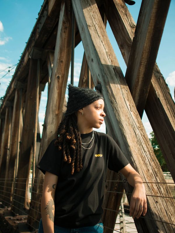 Person with dreadlocks wearing a black beanie and t-shirt, leaning on a wooden bridge.