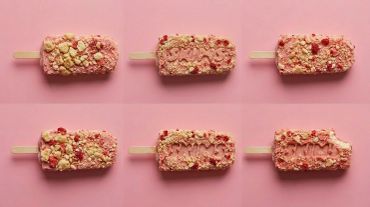Six pink popsicles with crumbs on a pink background, one partially eaten.