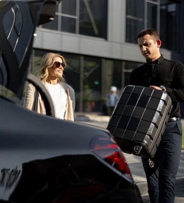 Man loads suitcase into car trunk as woman watches.