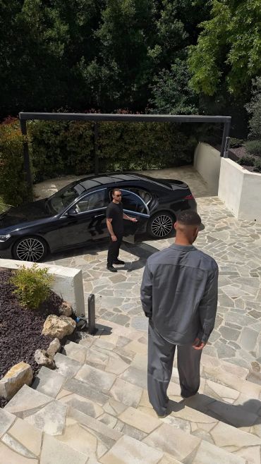 Two men near a black car in a driveway surrounded by greenery.