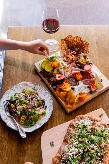 Charcuterie board, salad, flatbread, and red wine on table.