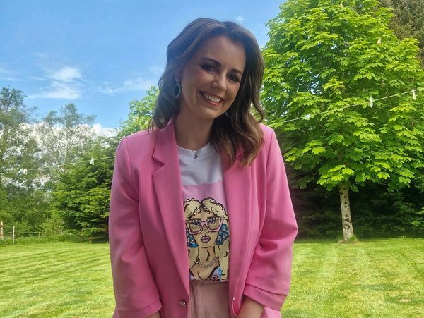 A smiling woman in a pink blazer stands outdoors with green trees and grass in the background.