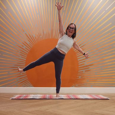 Woman balancing on one leg in front of an orange sunburst wall art.