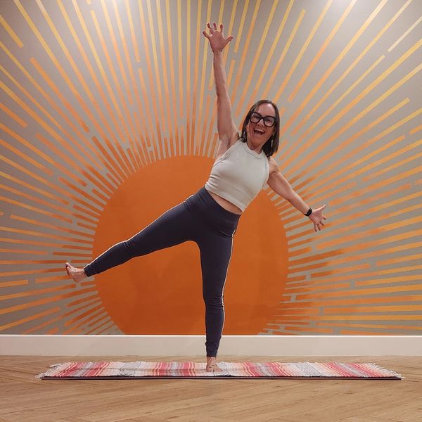 Woman balancing on one leg in front of an orange sunburst wall art.