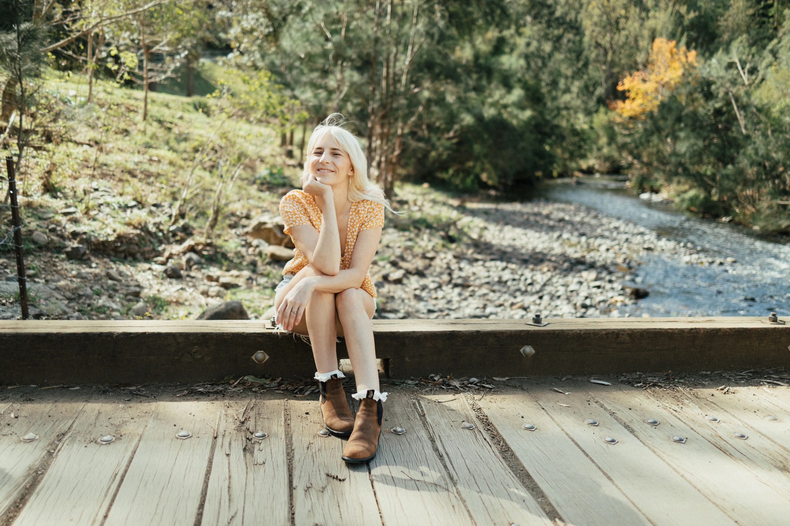 A woman in a yellow dress sits on a wooden bridge by a river.