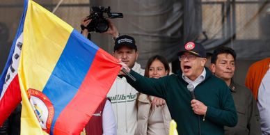 Man speaking at rally holding a Colombian flag with supporters behind him.