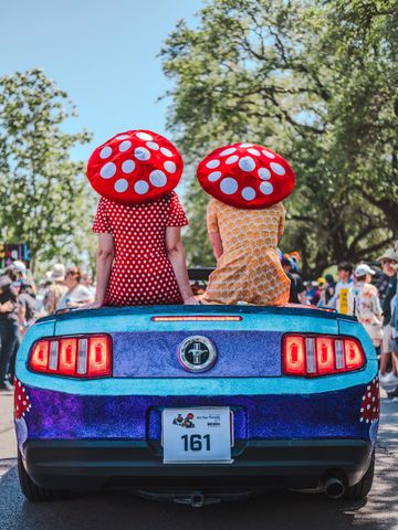 Two people wearing red polka dot mushroom hats sit on a blue car.