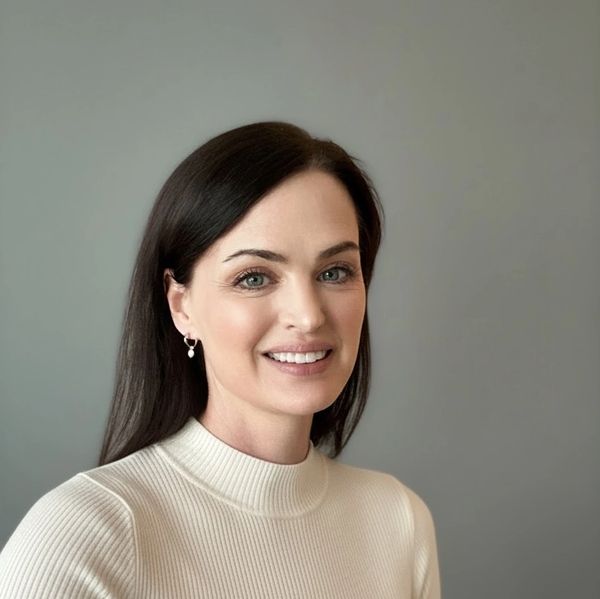 Smiling woman with dark hair wearing a cream sweater against a gray background.