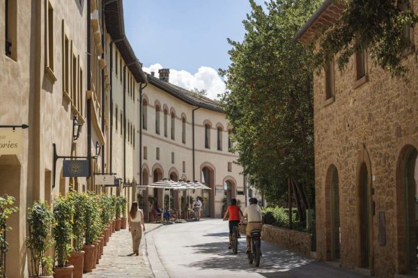 People walk and cycle on a quaint street with old buildings and greenery.