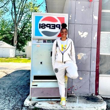 Smiling woman in white hoodie and sweatpants leaning on a vintage Pepsi vending machine.