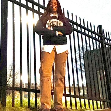 Person standing confidently in front of a tall metal fence during dusk.