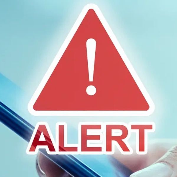 Photo of a mobile phone being held in a hand on a desk with a.red alert triangle on it
