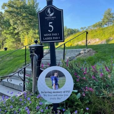 Memorial sign at golf course hole 5 with flowers and a photo tribute.