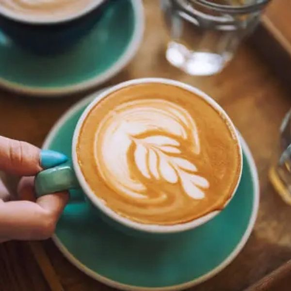 Hand holding a cup of latte with leaf latte art on a teal saucer.