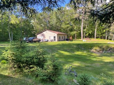House in a green wooded area with cars parked nearby.