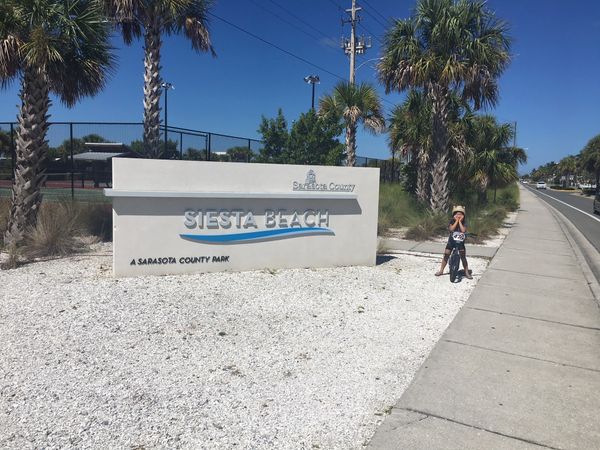 Siesta Beach sign in front of the tennis courts. Chloe sitting on her bike smiling at the camera