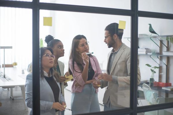 Three women staring at a man behind a window with sticky notes on it. 