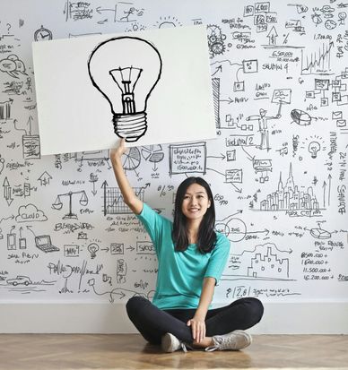 An asian millenial woman sitting criss-crossed while holding up a large picture of a light bulb. 