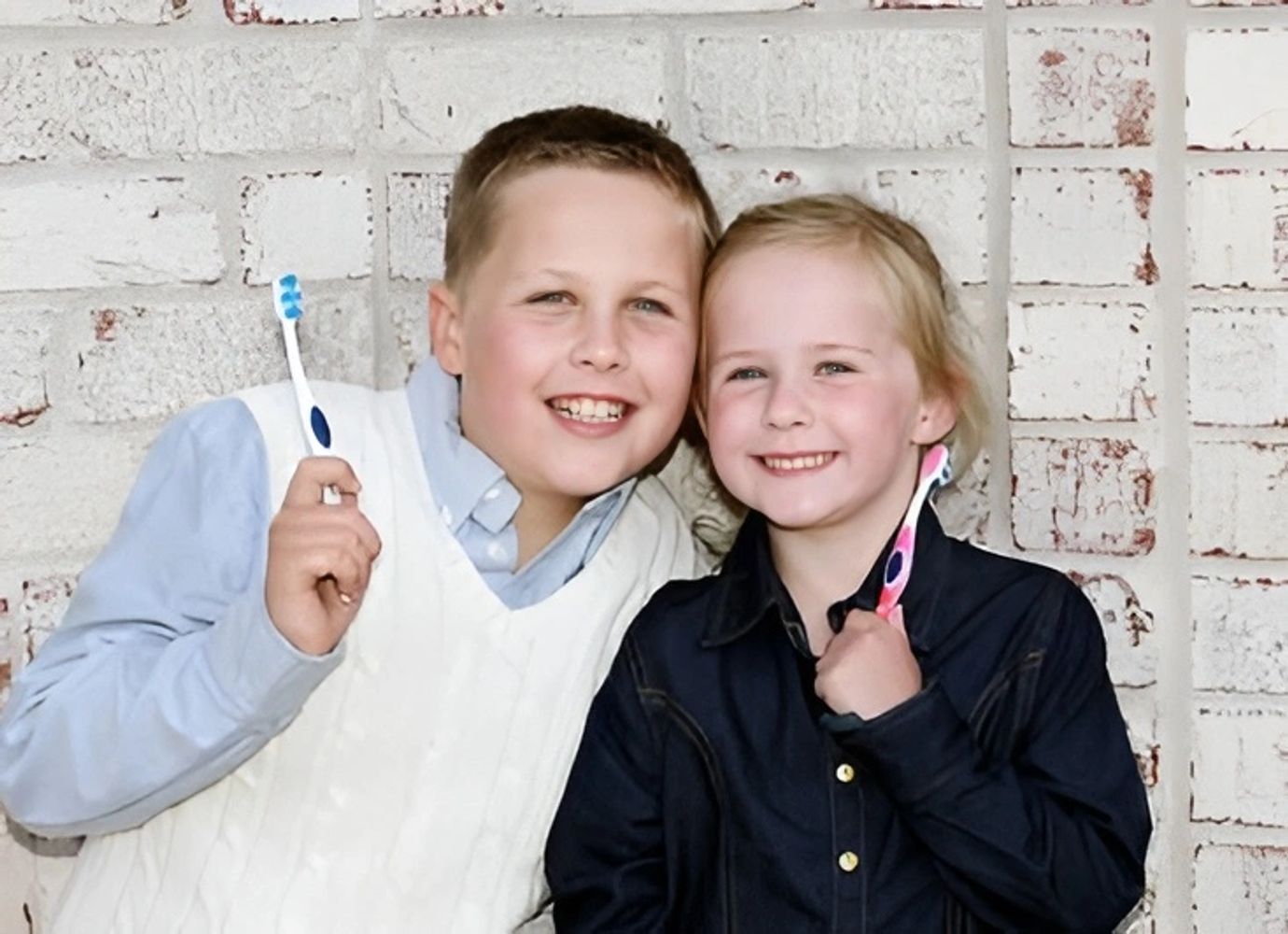 Two children stand against a white brick wall, each holding a toothbrush.
